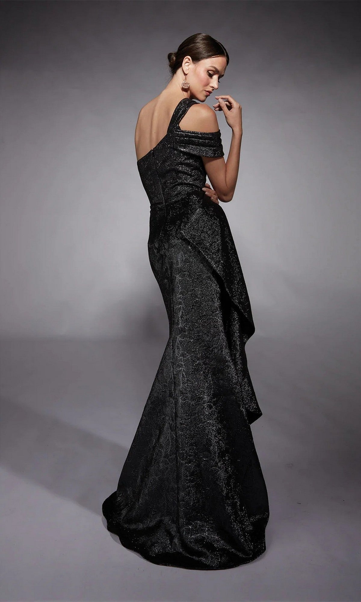 A woman poses with her back to the camera in the fitted, off-shoulder, textured black Alyce 27734 long formal dress against a plain gray background.